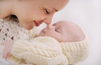A woman gently touches her nose to a sleeping baby, both wearing knitted cream clothing.
