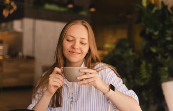 Portrait of gorgeous blonde smiling lady smelling, enjoying of coffee and drinking cappuccino from cup while resting in restaurant. Woman wear stripped shirt., 