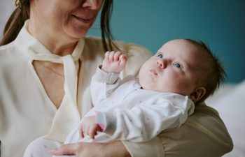 Woman holding newborn baby in vibrant beige room. Baby wearing white onesie, gazing upwards while mother lovingly holding infant close